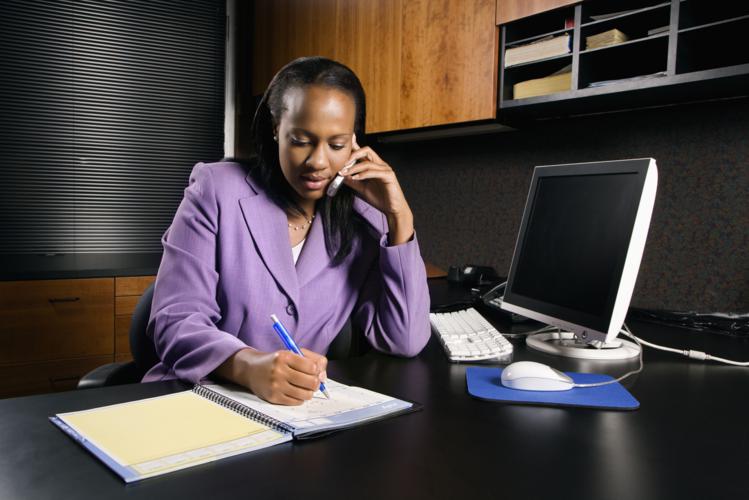 Woman working in office. African-American young adult business woman talking on cell phone and writing in planner in office.